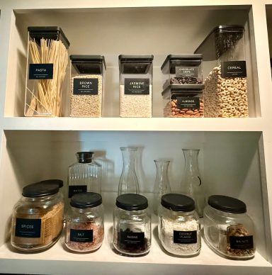 Assorted clear storage jars with labels, containing various dry ingredients and pasta.