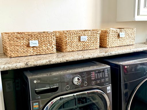 Three woven storage baskets on a granite countertop above a washing machine and dryer.